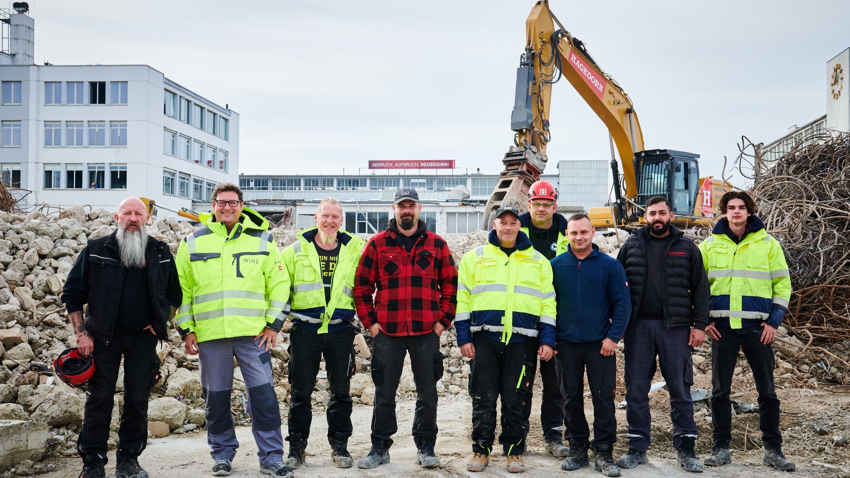 Vielfältiges Arbeiterteam steht lächelnd vor Baumaschinen auf einer Abbruchstelle.