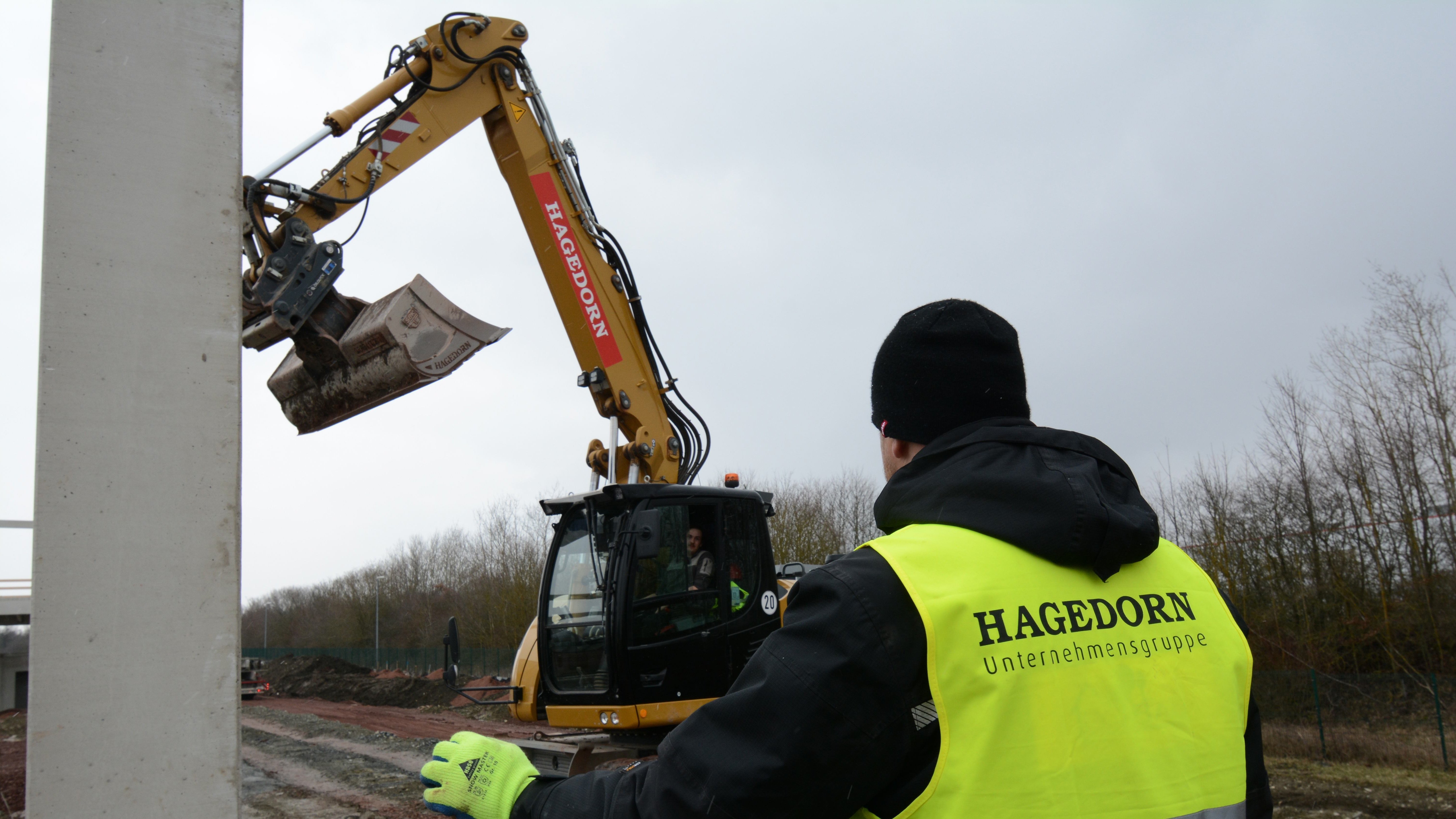 Arbeiter in Warnweste steuert Baggerarm auf Baustelle.